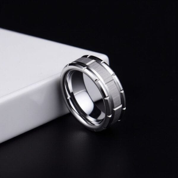 On a black minimalist background a ring rests against a white ledge. The focal point of the image is the silver tungsten ring. It has a brickwork pattern including some brushed silver sections.