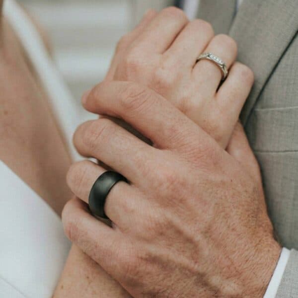 During the wedding ceremony, the couple turn to face each other. Holding hands together, the new wedding bands are visible. The woman is wearing a silver ring while the man is wearing a black tungsten ring with a slightly brushed finish.