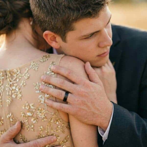 The man leans forward to embrace his partner. While she is dressed in a gold, he is wearing a navy suit. On his hand is a black tungsten ring that has a rose gold inside.