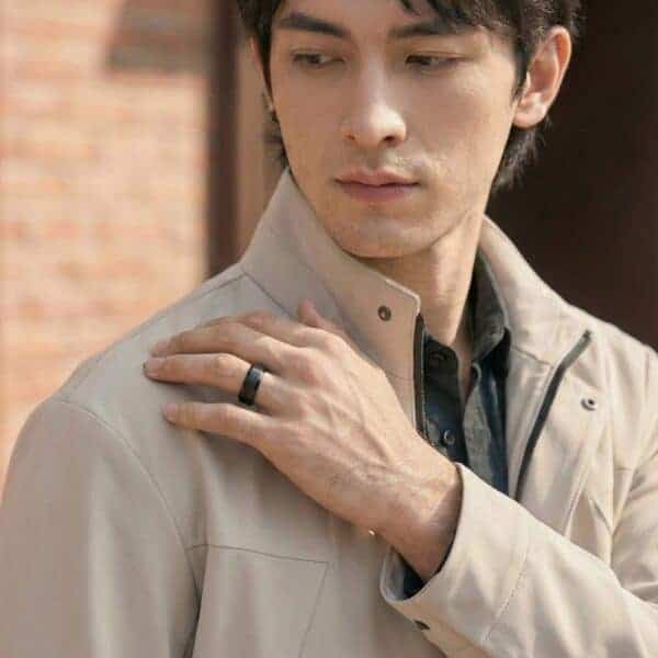 Wearing a raincoat, the man reaches his hand up to his shoulder. He wears a ring on his wedding finger. It is a domed black tungsten ring that has silver edges.