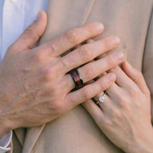 A zoomed in shot showing the hands of a couple. Holding their hands up to the mans jacket, the women has a silver diamond ring while the man has a black and red tungsten ring. The ring features an inlay of red carbon fibre. It looks like these are engagement rings.