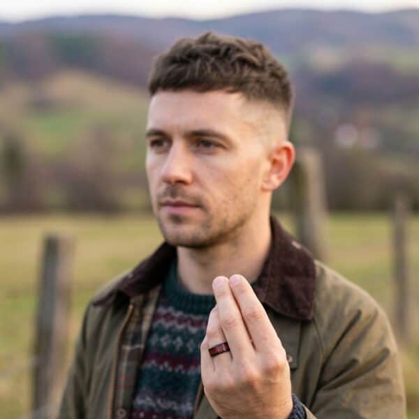 Out in the countryside, the image shows a man hiking. With a neutral expression on his face, he holds his hand up in front of him. He is wearing a black and red ring. The black is polished tungsten and the red is an inlay made of carbon fibre. The ring has stylish bevelled edges.