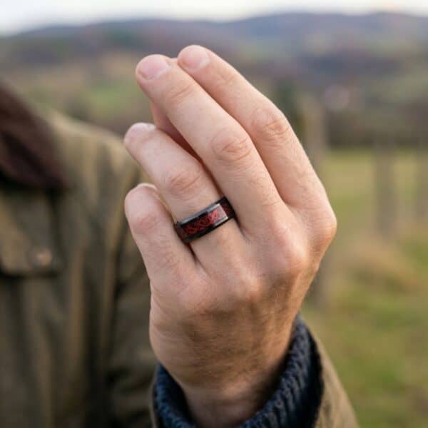 The camera is zoomed in on a mans hand. The man looks like he is out walking in the English countryside. He has a ring on his hand that is black and red. It is a black tungsten ring that has an inlay of red carbon fibre.