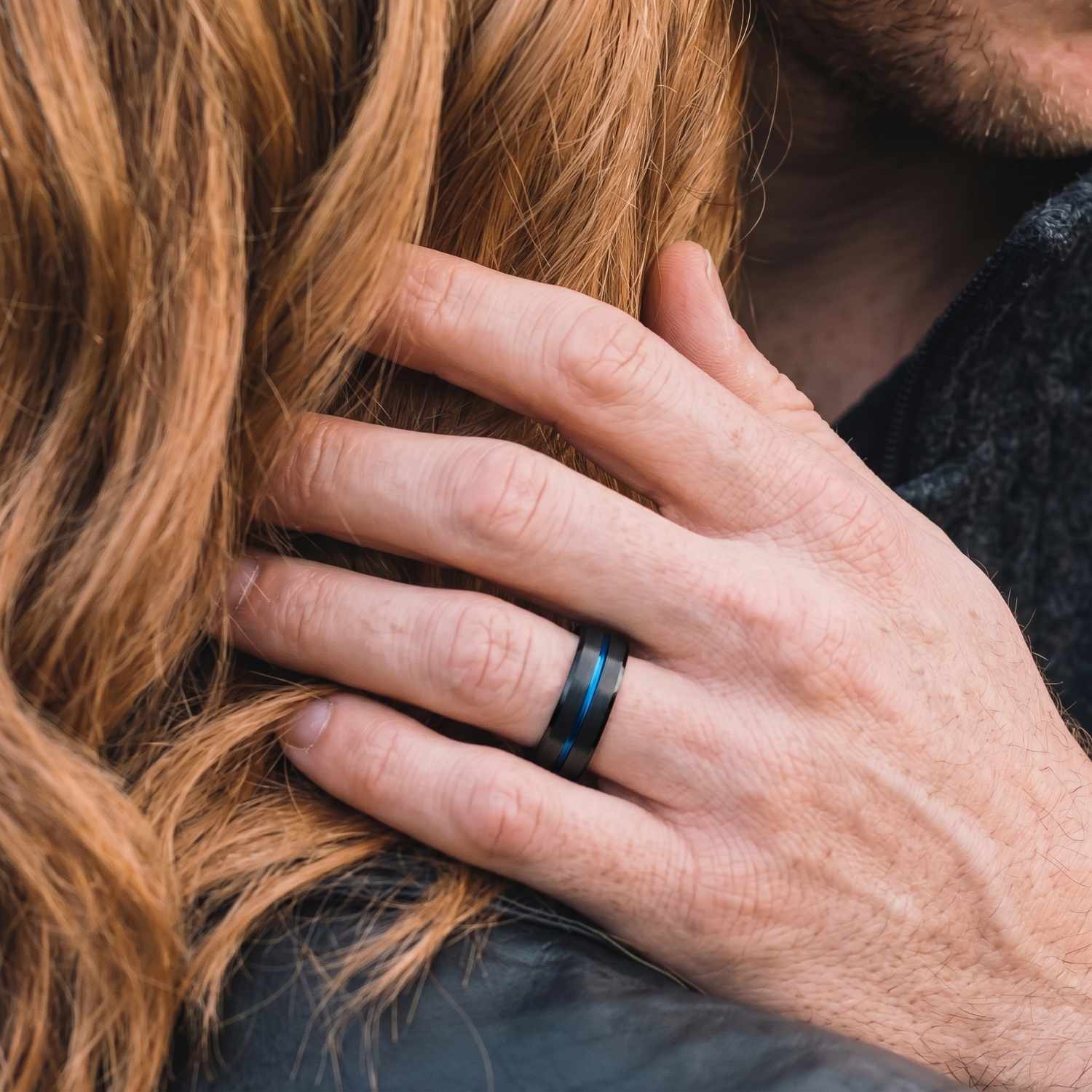 Close-up of a man's hand wearing the Unswerving black titanium ring with a polished blue groove as he gently touches his partner's hair.