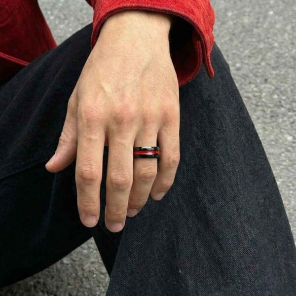 A man sits with his hand across his knee, showing his hand in close up. He is wearing a black tungsten ring that features a groove of vibrant red at the centre of the ring.