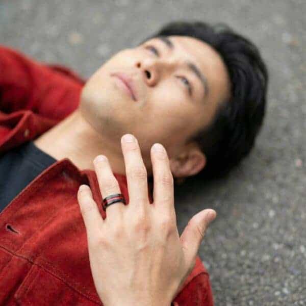 Laying on the ground, a man faces upwards with his hand stretched out. On his wedding finger is a black tungsten ring that has a groove of red running through the centre of the ring.