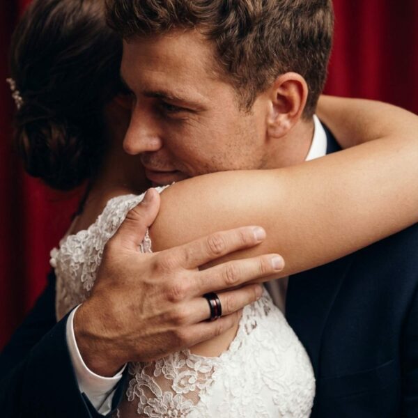 The wedding couple embrace. The mans hand is visible showing his black and red tungsten wedding ring.