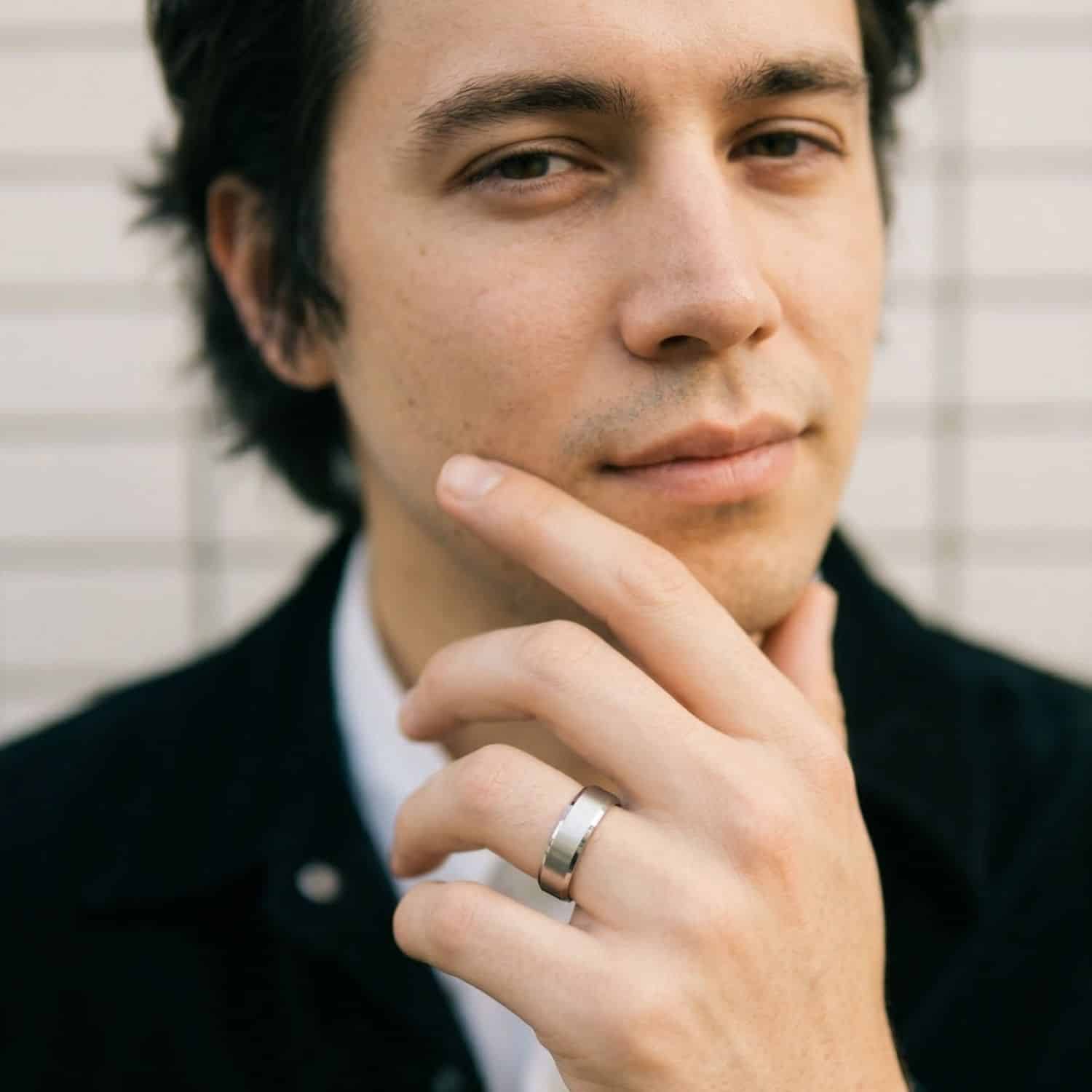 A man has a slight smile of his face as he looks into the camera. He has his chin resting on his hands. On his hand he is wearing a silver titanium ring. It has a brushed centre and polished bevelled edges. The inside of the ring is blue.