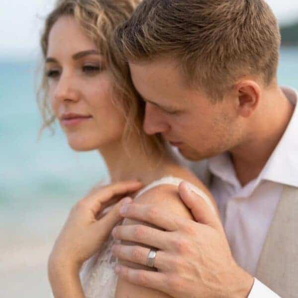 A wedding photograph of the happy couple as they celebrate their special day on the beach. The man embraces his new bridge while she looks off to one side with a smile of her face. The man has his hand on the woman's shoulder. You can see his wedding ring. It is a silver titanium ring - it has a brushed centre and polished bevelled edges. The interior of the ring is a vibrant blue.