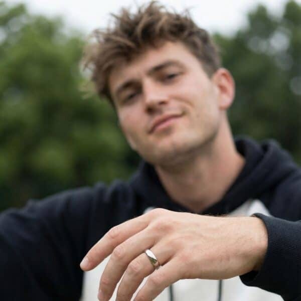 Smiling into the camera a man smiles as he relaxes in parklands. He is wearing a black and white hoodie and has a polished silver ring on his hand. The ring is slightly domed and looks like a wedding band. It is made with titanium.