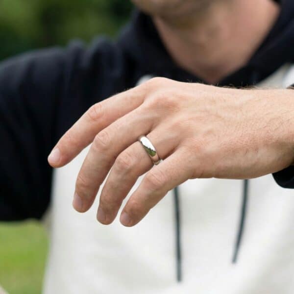 A zoomed in photo of a man wearing a white and black top. There is greenery in the background. The mans hand is at the front of the photo. He is wearing a silver titanium ring.