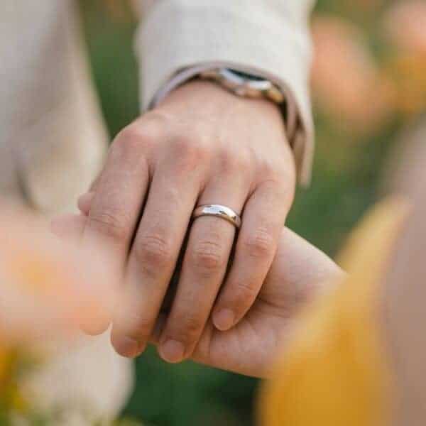A zoomed in shot of a married couple holding hands. Blurred flowers frame the shot, with the mans ring prominent on his hand. It is a polished silver titanium ring.