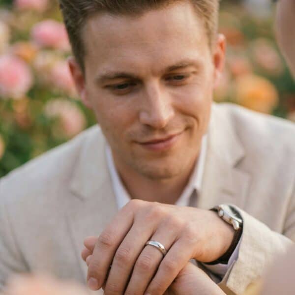 Newly married, the image focuses on the newly married man. He is holding the hand of his partner but they are just out of view of the photo. The man is smiling and has his hand on his partners hand. He is wearing a shiny silver titanium ring.