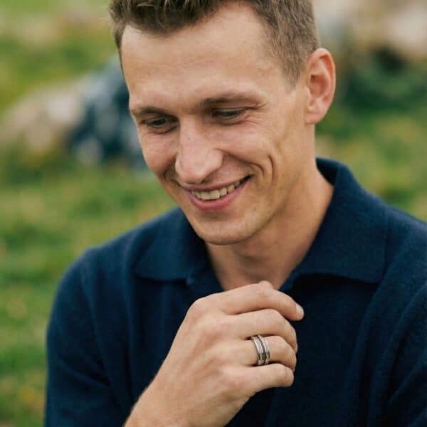 With a backdrop of green grass, a man sits smiling. He is showing his hand, wearing a silver tungsten ring that has an inlay of wood and two inlays of antler, on either side of the wood.