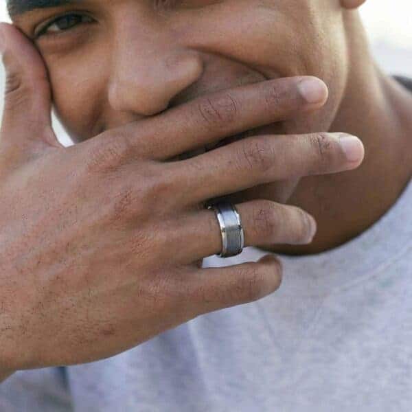 A man faces the camera with a wide smile on his face. He is covering his mouth with his hand. On his hand he wears a silver tungsten ring. It has a brushed silver centre section and polished edges.