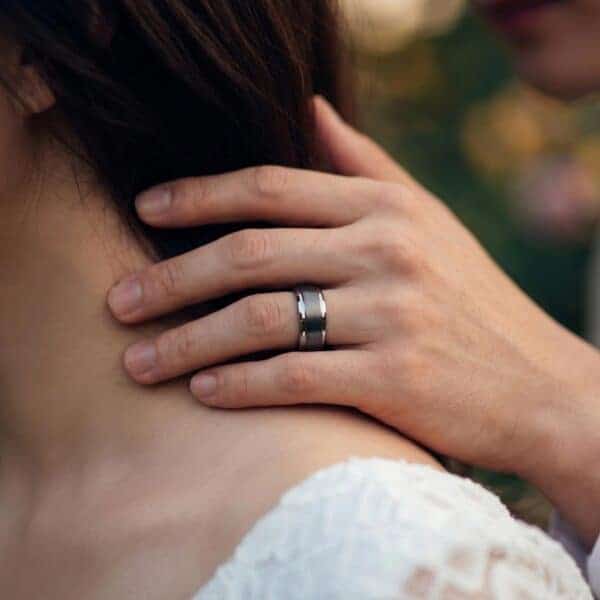 A close up shot of a newly married couple on their wedding day. The man rests his hands on the brides shoulder. You can see his wedding band - a silver tungsten wedding ring that features a brushed centre section and polished edges.