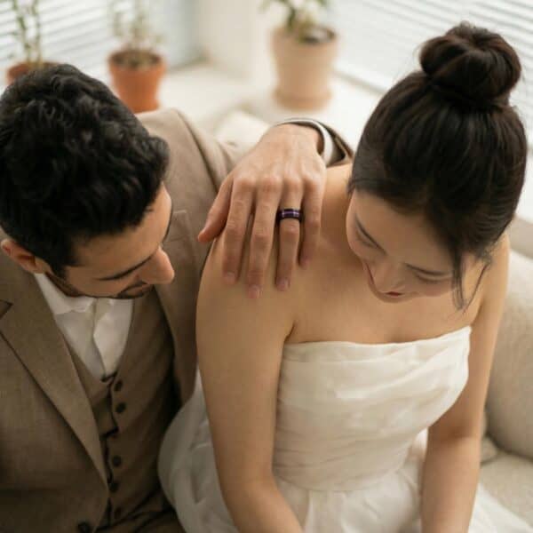 On the wedding day, the photographer takes a natural photograph of the bride and groom. Sharing a quiet moment together, the couple sit together with the man resting his hands on his partners shoulder. The bride wears a white wedding gown and the man wears a brown suit. The man is wearing a black tungsten ring which features a recessed groove of purple, and a purple interior.
