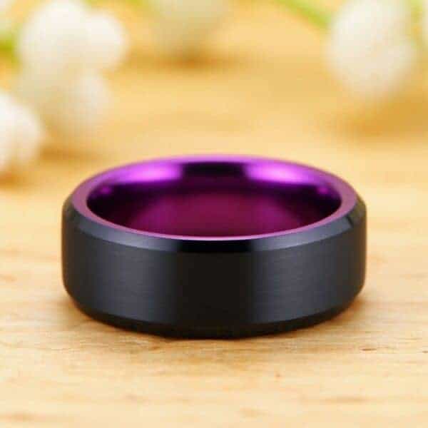 On a light wood surface and with white flowers scattered in the background, a ring lays on its side at the front of the image. The ring is a black tungsten ring with a vivid purple interior.