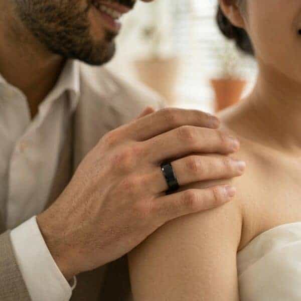 On their wedding day, the man places his hand on the shoulder of his bride. Wearing a white shirt and beige blazer, he is wearing a black tungsten ring. It has a brushed centre section and polished bevelled edges. The inside of the ring is polished purple.