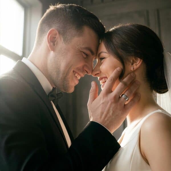 A happy wedding couple stand opposite each other looking into the face of one another. The woman wears a white wedding dress and the man is dressed in a black suit. The man has his face held to his brides face as they smile happily together. On his hand, the man is wearing a silver wedding band. It is a tungsten ring that features raised brushed sections and a groove of polished silver, as well as silver edges.