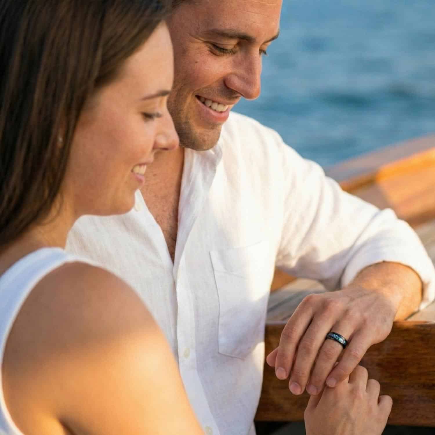 A couple sit together on a boat. They look towards the ring on the mans hand. It is a black titanium ring with an abalone shell inlay.