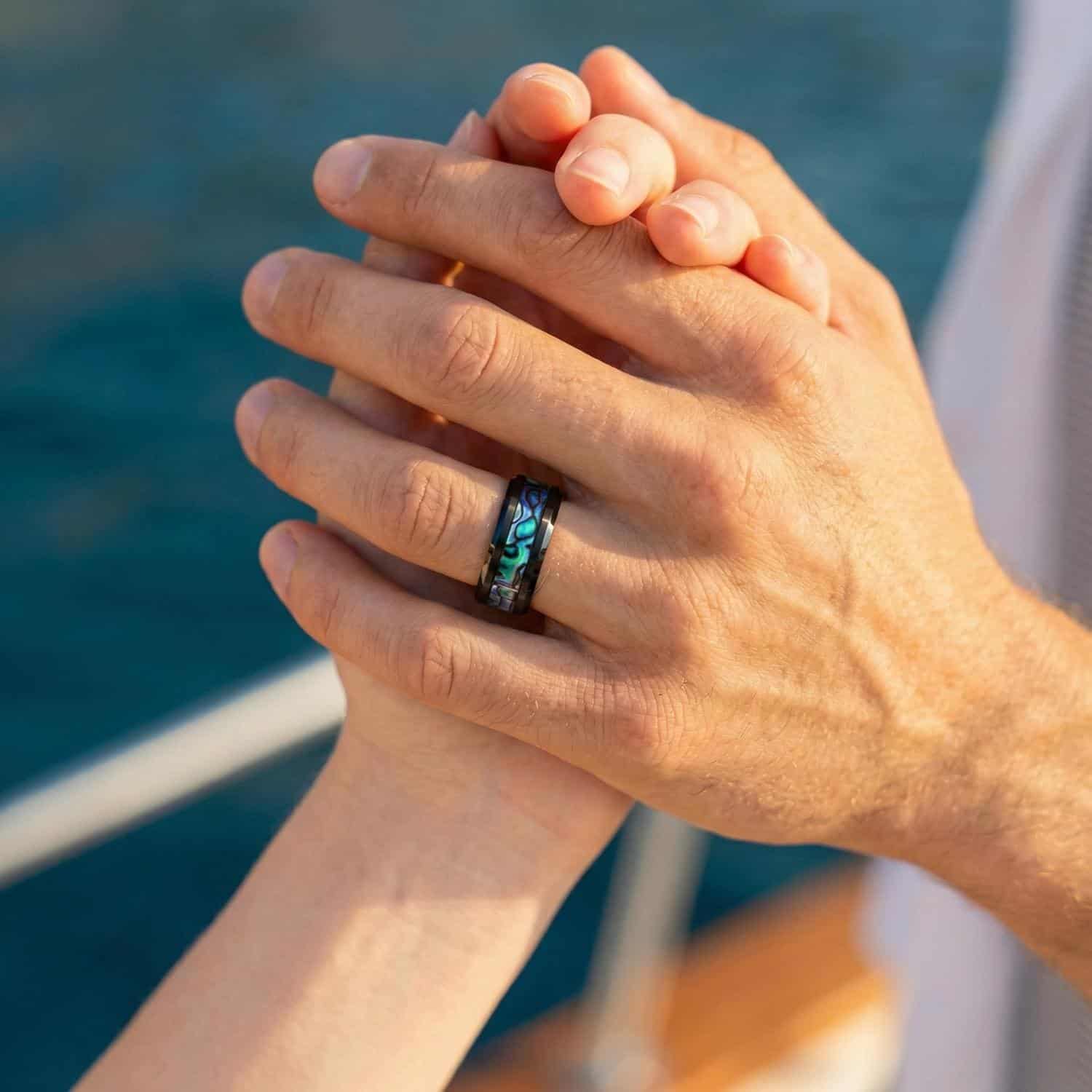 On board a boat, a couple hold hands. This is a close up of the hands. On the mans hand is a black titanium ring with a striking inlay of abalone shell.