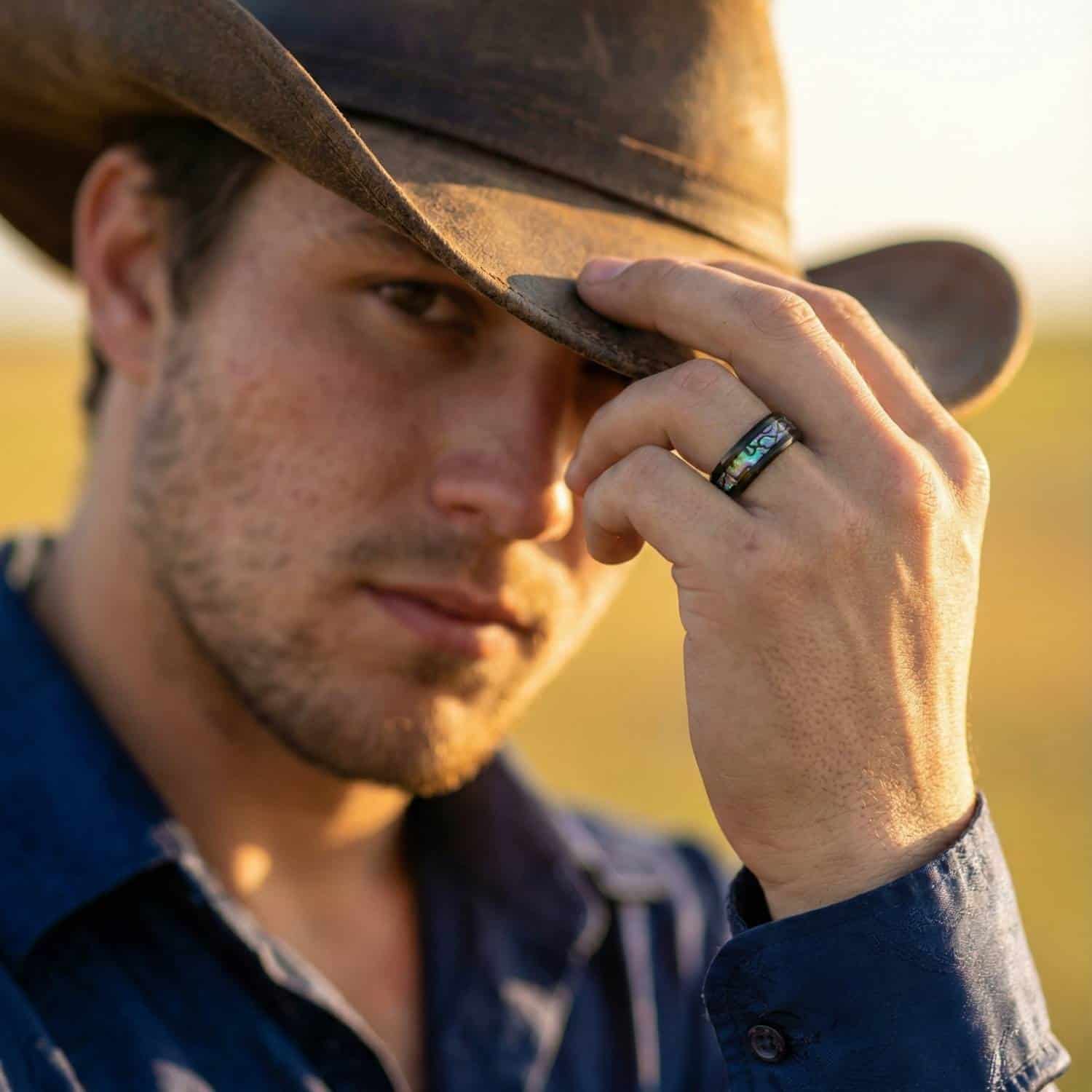 A man looks towards the camera wearing a cowboy hat. As he holds the rim of the hat, his ring is clearly visible. It is a black titanium ring with an inlay of abalone shell.