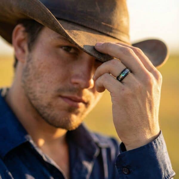 A man looks towards the camera wearing a cowboy hat. As he holds the rim of the hat, his ring is clearly visible. It is a black titanium ring with an inlay of abalone shell.