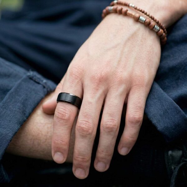 A close up of a mans hand wearing a black tungsten ring on his index finger. The ring has a brushed black centre section and polished black edges. The man is wearing beaded bracelets.