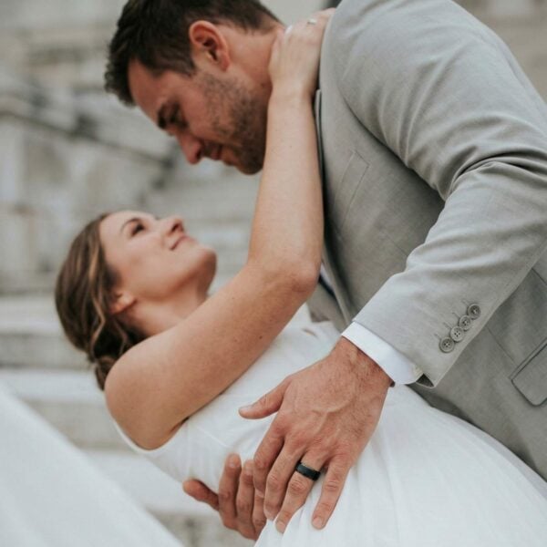 The newly married couple dance together. The bride leans back as the man supports her with his arms around her waist. He is wearing a black tungsten ring. The centre of the ring is brushed and the sides are polished.