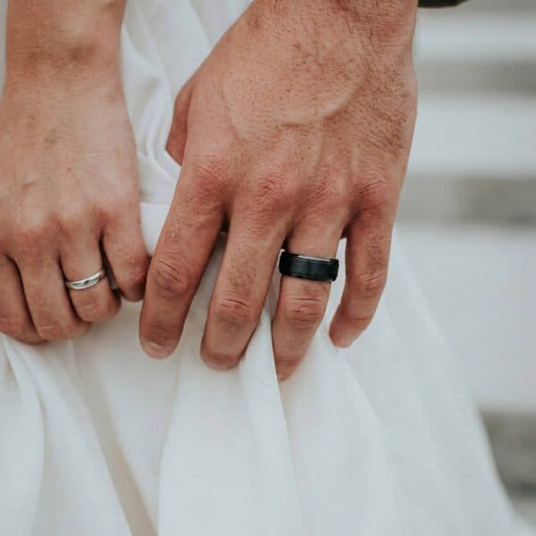The 'just married' couple gently touch their hands togethr. Against the white of the brides wedding dress, you can see the two new wedding rings. The woman is wearing a silver band while the man is wearing a black tungsten ring. It has a brushed section at the centre of the ring and polished bevelled edges.