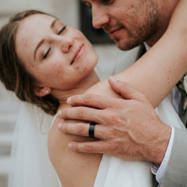 The groom leans forward to kiss his bride. She is smiling and has the side of her face towards him. In their embrace, you can see the mans wedding band. It is a black tungsten ring. It has a brushed centre section and polished edges.