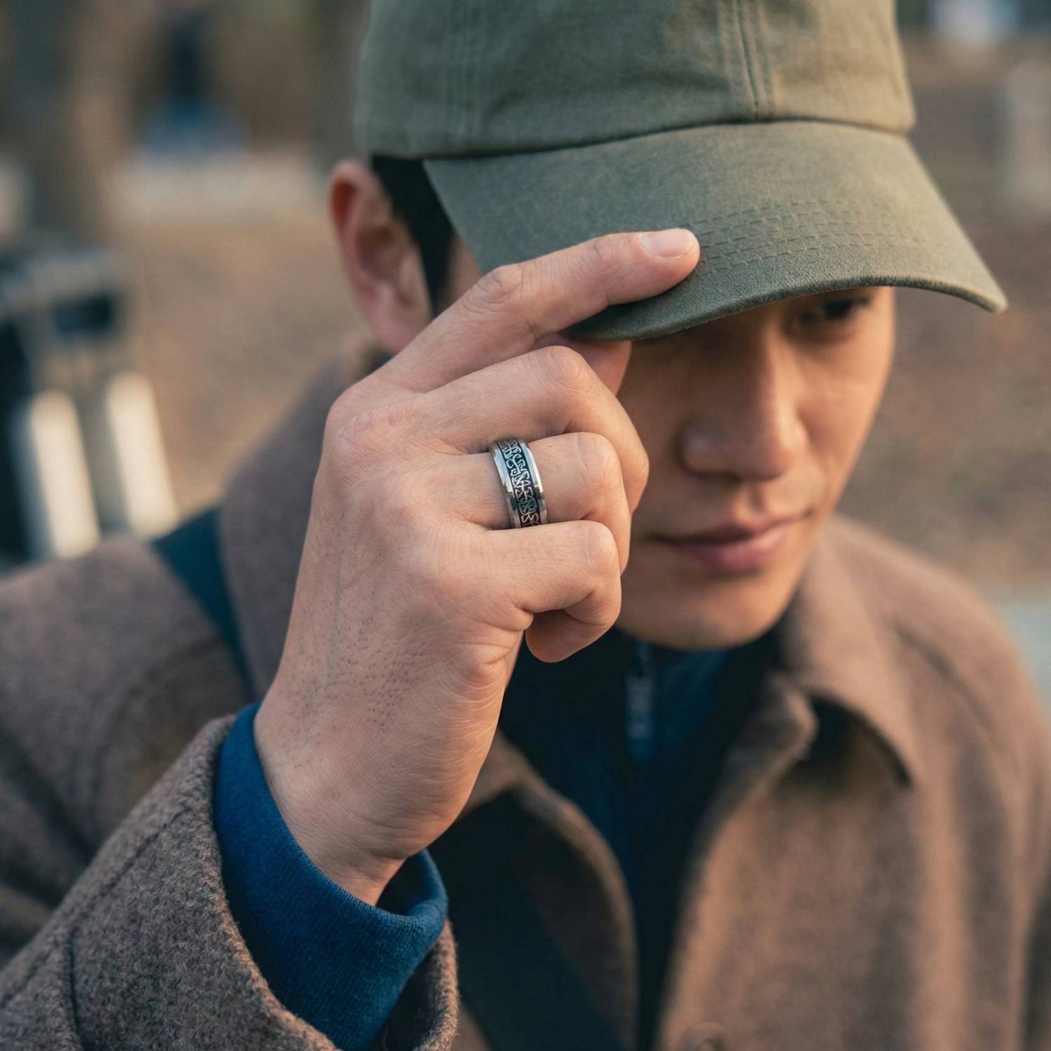 A man holds his hand up to adjust his baseball cap. On his hand he wears a silver tungsten ring. It has an inlay of carbon fibre in an intricate celtic knot pattern. He wears a brown overcoat.