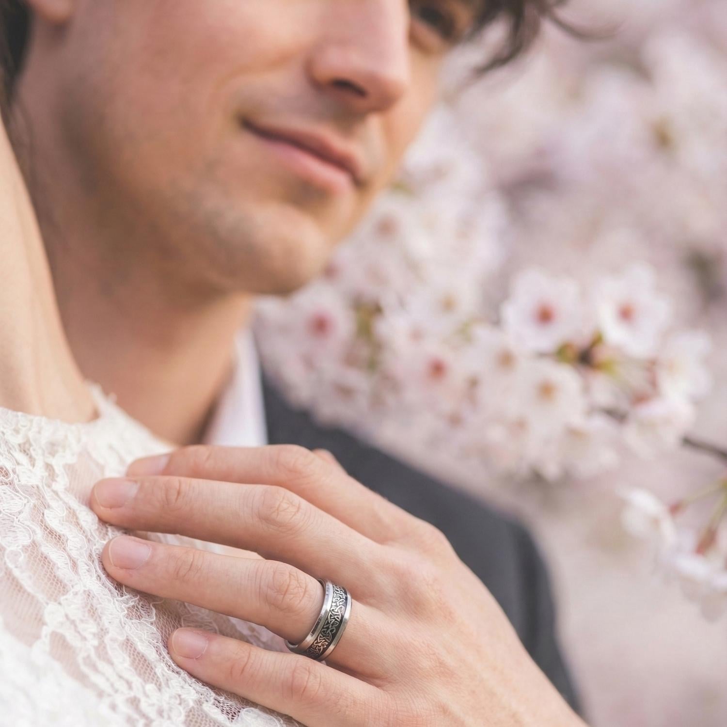 With a blossom tree behind them, the wedding couple embrace. There is a close up view of the mans ring - it is a silver tungsten ring with an inlay of black carbon fibre with an intricate Celtic knot pattern.