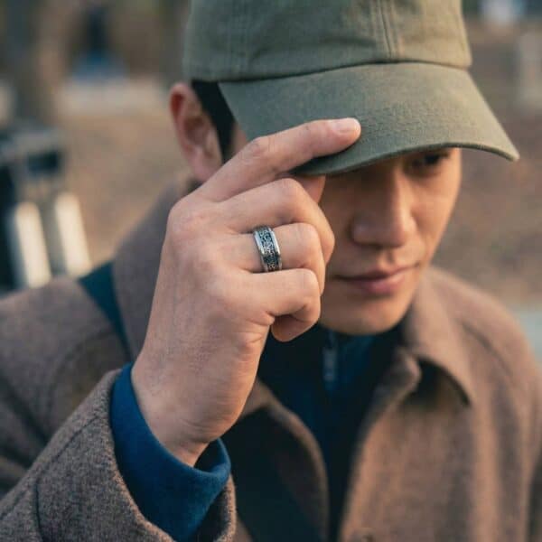 A man holds his hand up to adjust his baseball cap. On his hand he wears a silver tungsten ring. It has an inlay of carbon fibre in an intricate celtic knot pattern. He wears a brown overcoat.