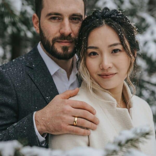 A couple stand together in the snow, facing the camera. The man rests his hand on his partner's shoulder. On his wedding finger is a gold titanium ring.