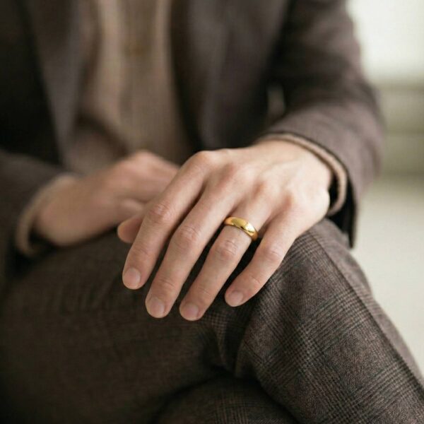 Wearing a brown suit, a man sits with his legs crossed. He rests his hand on his knee. On his hand, he is wearing a gold titanium ring.