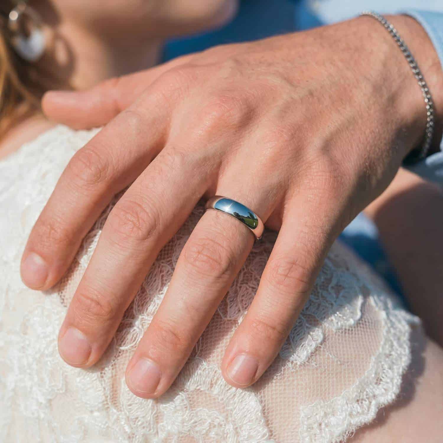 A close of photo of a newly married couple. The man has his hand resting on the brides shoulder. The camera has zoomed in on the mans hand. He is wearing his new wedding band which is a slim style polished silver titanium ring.
