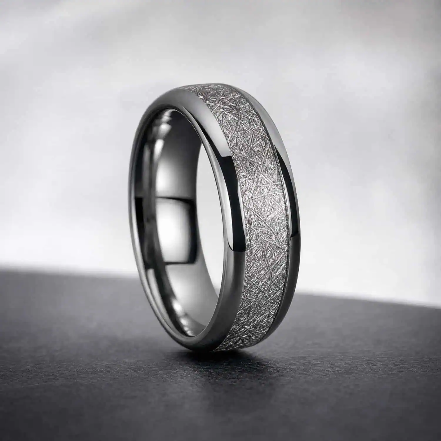 Silver tungsten ring standing upright on a dark grey surface against a light grey minimalist background, showcasing its faux meteorite centre inlay and polished finish.