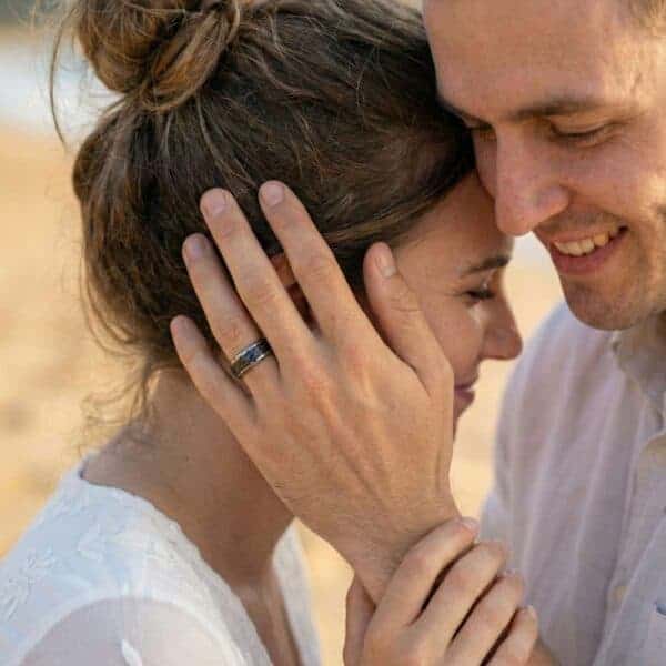 Standing close together, a couple smile with happiness. The man holds his hand to her cheek. He is wearing a silver tungsten ring that has a black centre made of faux meteorite.