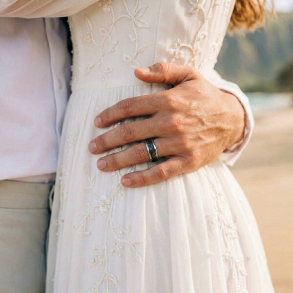 At the wedding ceremony, the bride and groom come together for an embrace. The bride is wearing a traditional white wedding gown. With his hand around her waist, you can see the man's wedding ring. It is a silver tungsten ring that has a black faux meteorite centre.