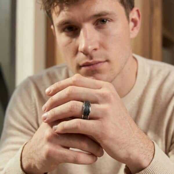 Looking into the camera, a young man looks thoughtful. With his hands held in front of him, he is wearing a silver tungsten ring. The centre is a black faux meteorite.