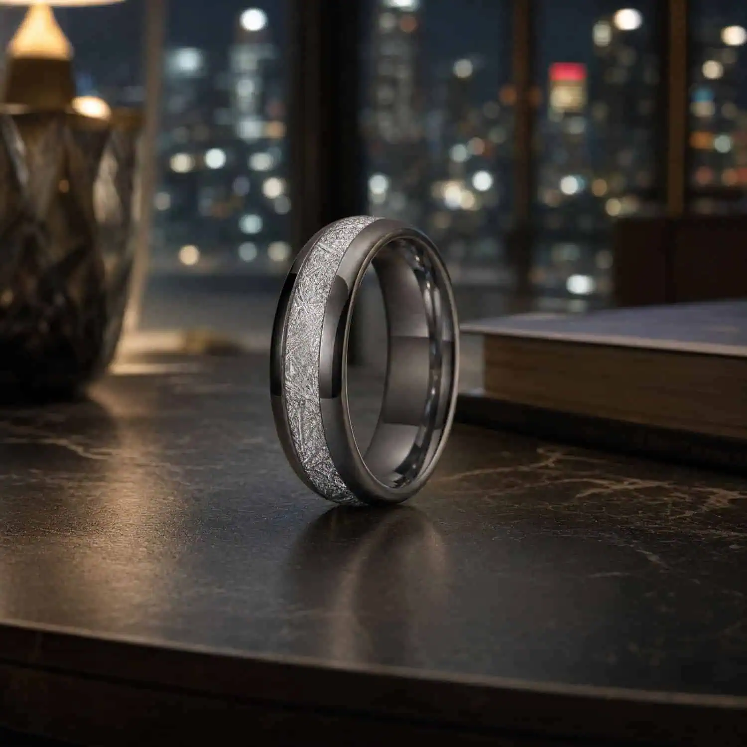 Men's silver tungsten ring with a meteorite-style inlay standing on a modern coffee table beside a closed book, with illuminated city skyscrapers visible through a large window.