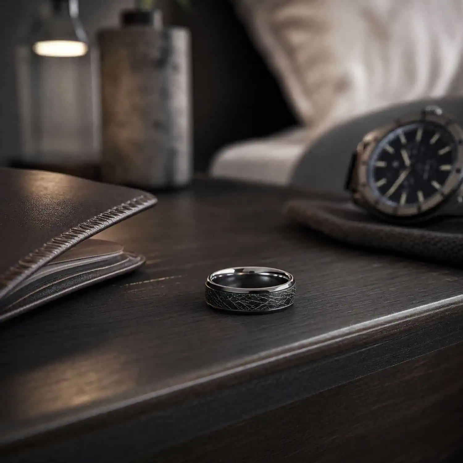 Men's polished silver tungsten ring with a black faux meteorite inlay resting on a bedside table beside a black watch, lamp and leather wallet.