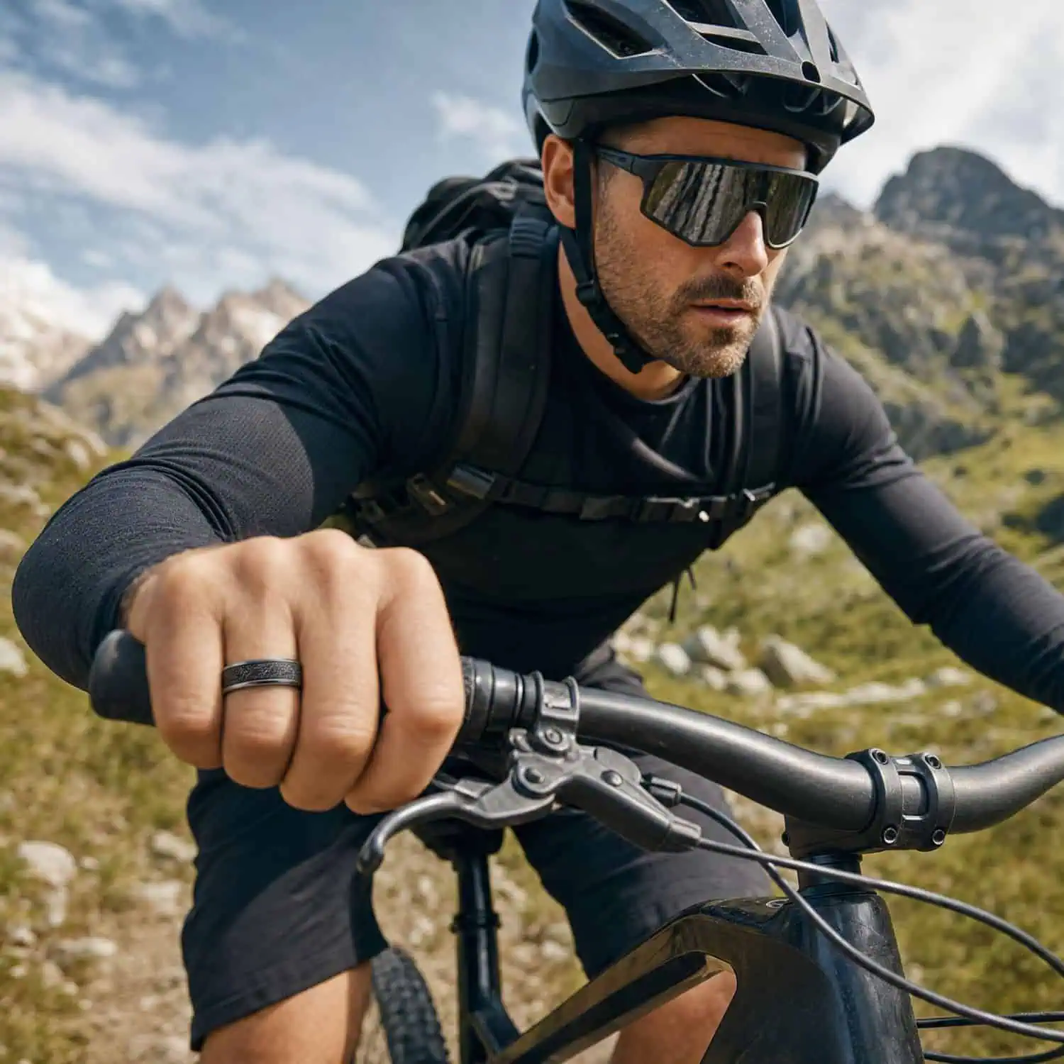 Man mountain biking along a gravel trail beneath sunny skies while wearing a silver tungsten ring with a black faux meteorite inlay, with mountain peaks in the background.
