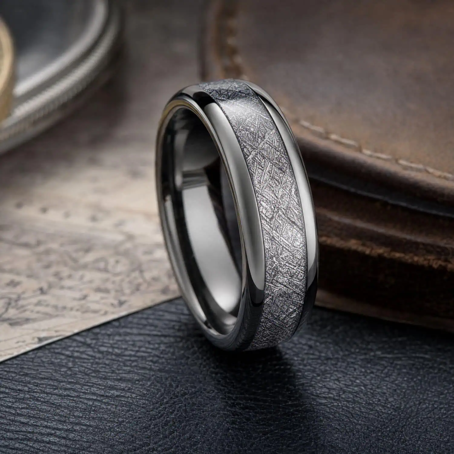 Men's polished silver tungsten ring with a faux meteorite centre inlay standing upright beside a travel map and leather bag.