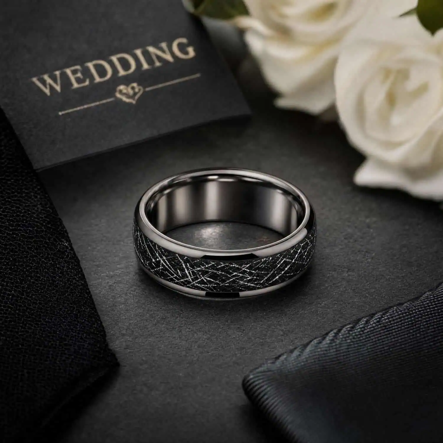 Men's silver tungsten wedding band with a black meteorite-style inlay displayed beside white wedding flowers, a dark tie and wedding order of service.