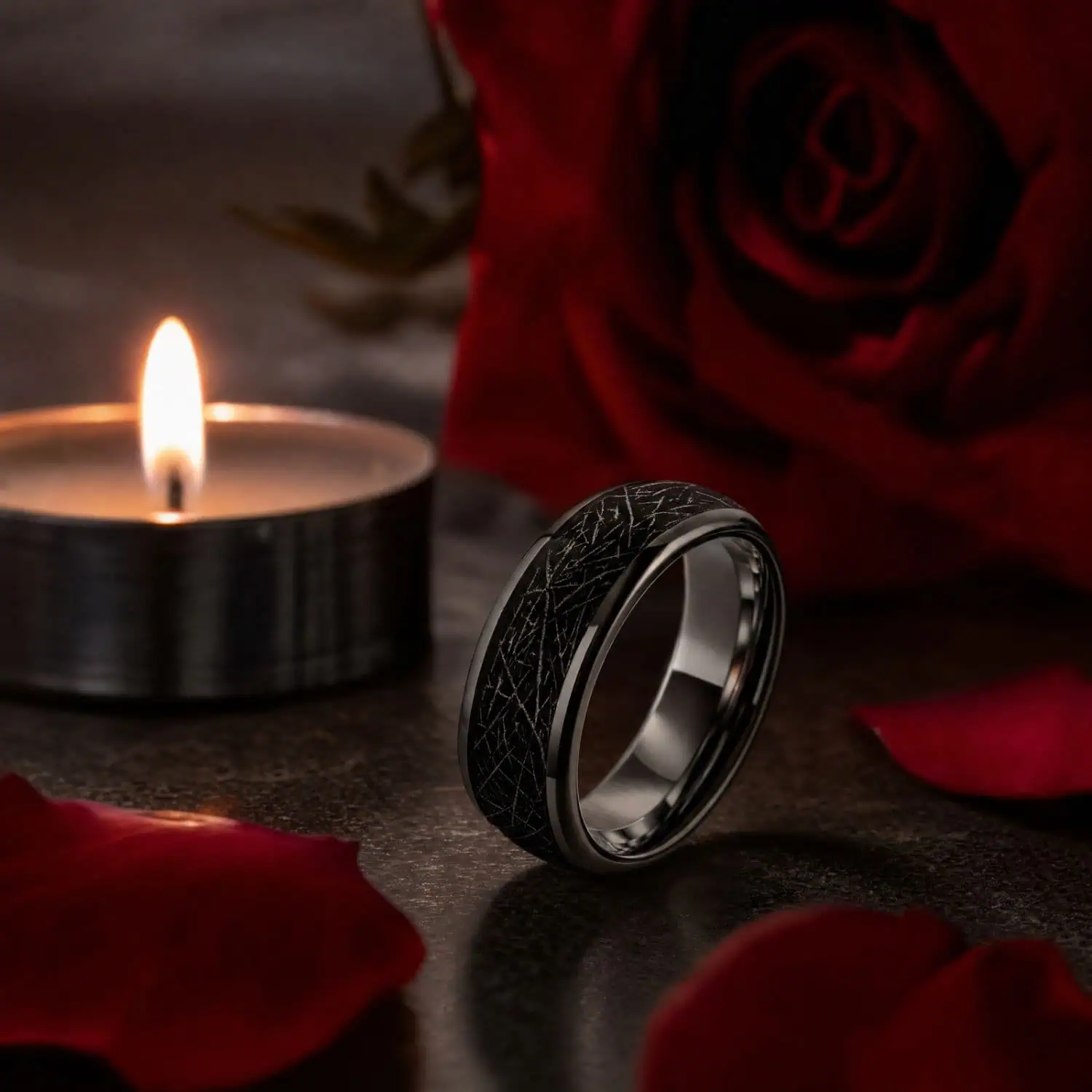 Meteorite-style tungsten wedding band standing upright beside a glowing tea light candle, red rose and scattered rose petals.