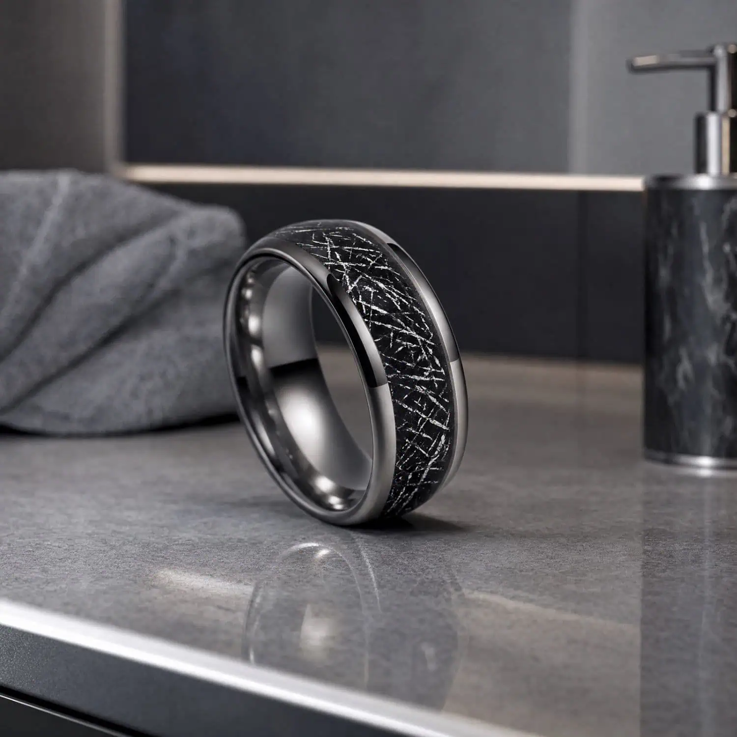 Polished silver tungsten ring with a black meteorite-style centre standing upright on a reflective bathroom countertop beside a grey marble soap dispenser.