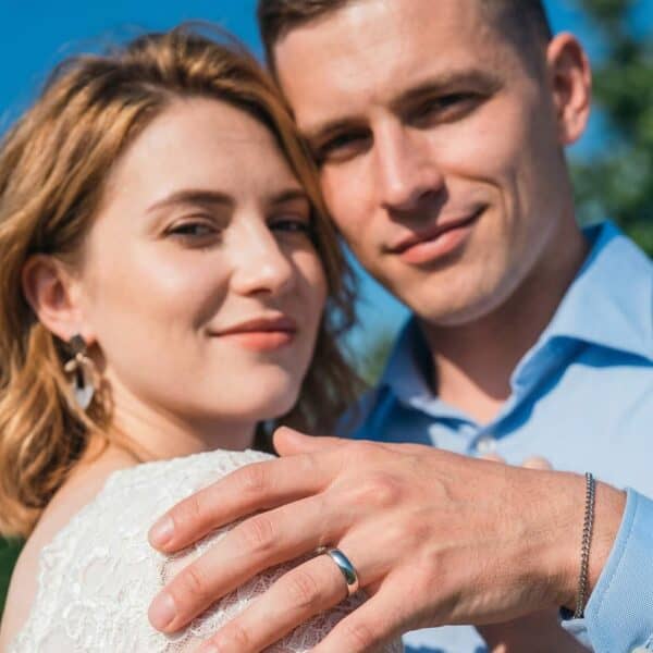 On their wedding day, a couple pose for another wedding photograph. Smiling into the camera lens, the couple are standing opposite each other and are loosely embracing. The man has his hand resting on the brides shoulder. He is wearing a slim style titanium wedding band in polished silver